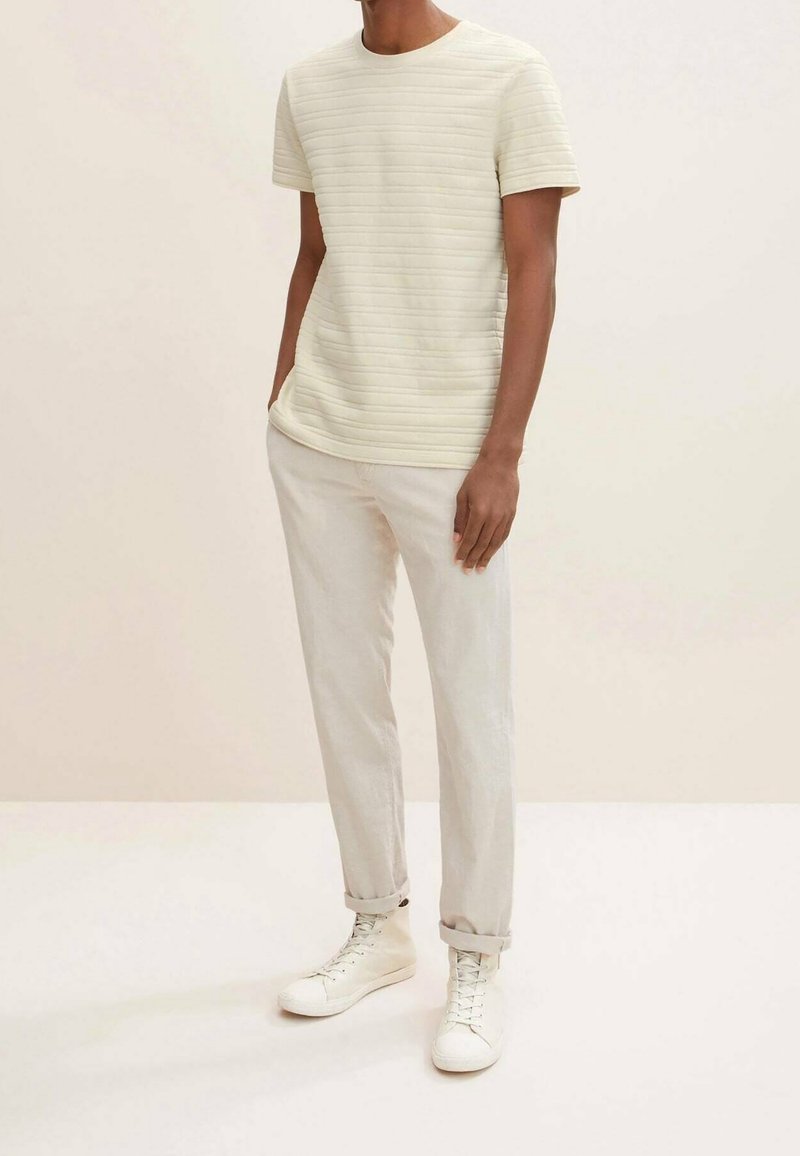 Homme portant une chemise à manches courtes texturée blanc cassé, un pantalon beige clair retroussé et des baskets montantes blanches, debout les mains dans les poches.
