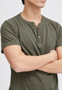 Grønn henley t-skjorte laget av myk, teksturert stoff. Har rund halslinning og tre treknapper foran. Korte ermer.