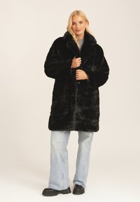 Black faux fur coat with a large collar, button front, and side pockets. Worn over light blue wide-leg jeans and black chunky shoes.