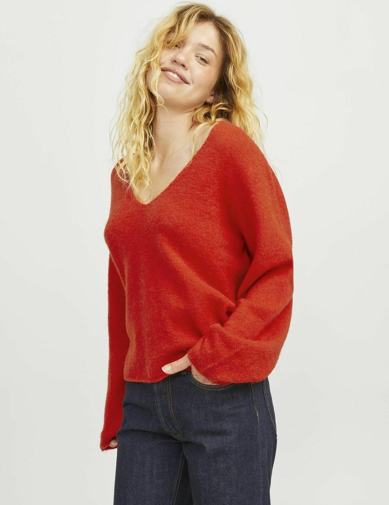 Roter V-Ausschnitt-Pullover aus einem weichen, strukturierten Stoff mit langen, lockeren Ärmeln. Kombiniert mit dunkelblauen Jeans. Einfaches, lässiges Design.