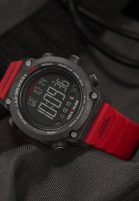 Digitale Timex Triathlon-Uhr mit schwarzem Gehäuse, rotem Gummiarmband und großem LCD-Display, das die Uhrzeit, Rundenzeit und Wasserbeständigkeitsindikatoren anzeigt.