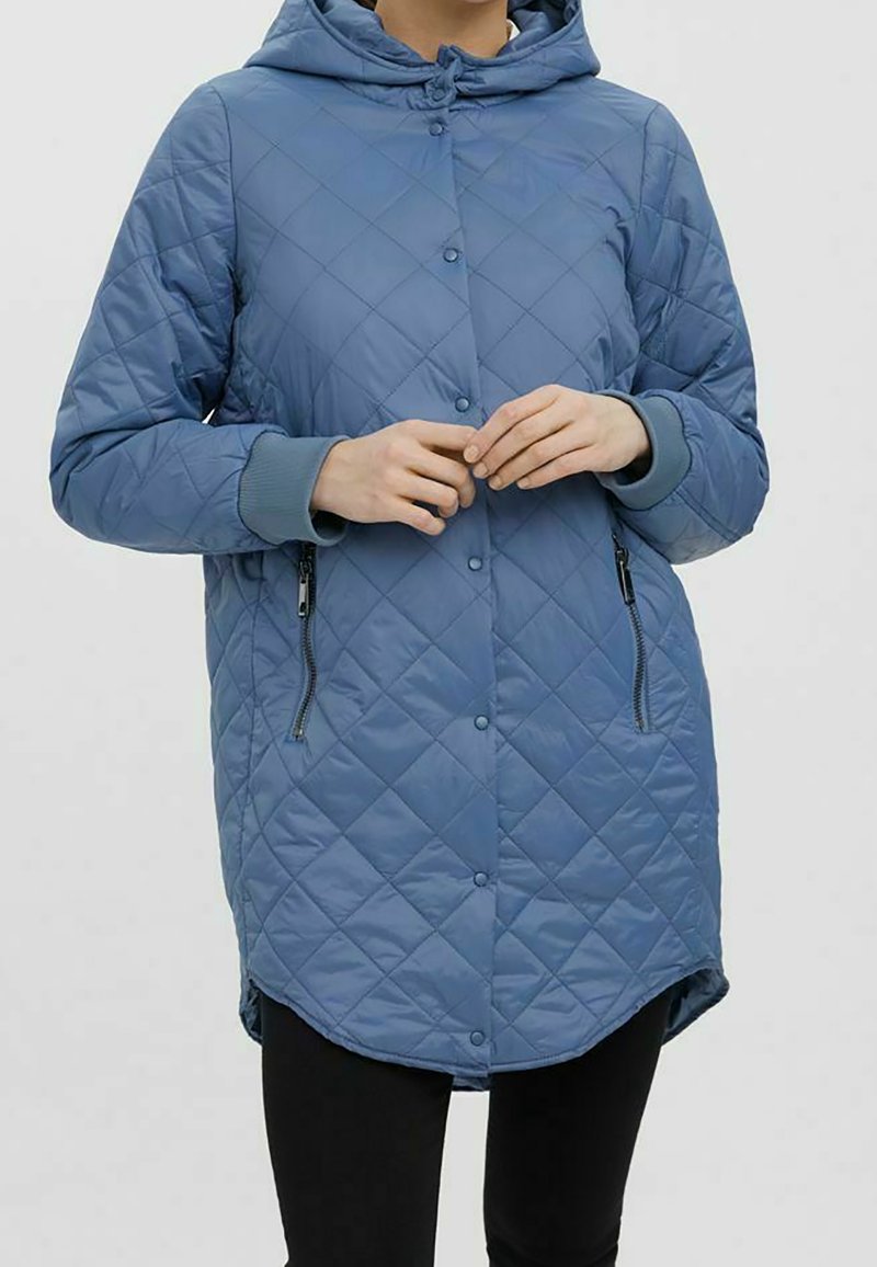 Femme portant une veste matelassée bleue à capuche avec boutons-pression et poches zippées, debout les mains jointes.