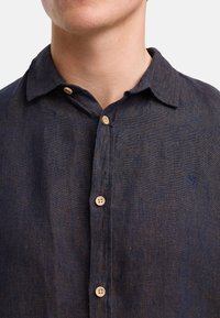 Camicia di lino blu navy con colletto button-down, caratterizzata da bottoni in legno e un piccolo logo ricamato nell'angolo in alto a sinistra.