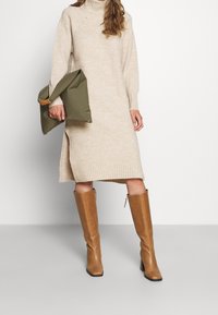 Beige ribbed turtleneck mekko, jossa on sivurei'ät, yhdistettynä ruskeisiin polvipitkiin saappaisiin ja vihreään laukkuun, jossa on nahkakorostus.