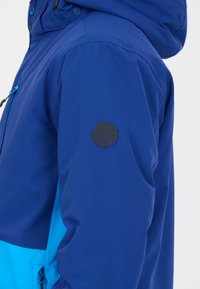 Blå och ljusblå vattentät jacka med texturerad yta, utrustad med en svart logotyp på vänster ärm och en justerbar huva.