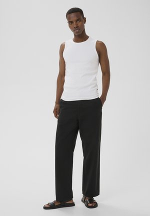 Homme debout les mains dans les poches portant un débardeur blanc, un pantalon noir à jambes larges et des sandales noires sur un fond uni.