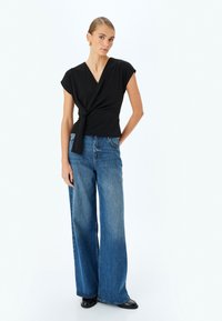 Top negro con mangas cortas, atado en la cintura, combinado con jeans de pierna ancha en azul deslavado y botas negras hasta el tobillo. Acabado de tejido suave.