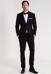 Zwart smoking met satijnen revers, wit overhemd en vlinderdas. Getailleerd ontwerp, gestructureerde stof en zwarte leren schoenen. Minimalistisch wit pochet.
