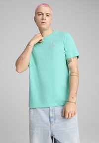 Mátově zelené bavlněné tričko s krátkým rukávem, na kterém je vyšitý malý logotyp Puma na hrudi. Nosené s světle modrými džíny.