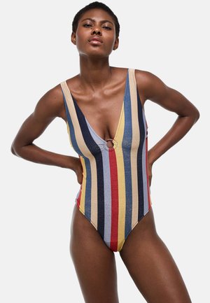 Femme portant un maillot de bain une pièce rayé vertical multicolore avec un décolleté profond et une petite découpe circulaire au niveau de la poitrine.