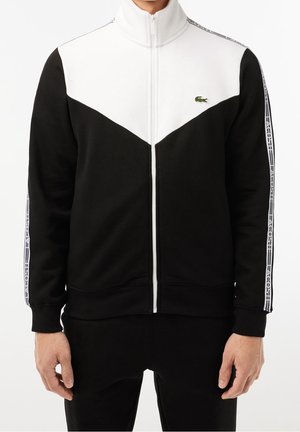 Zwart-wit jack met rits en hoge kraag, voorzien van een chevronpatroon, Lacoste-logo en gestreepte accenten op de mouwen. Zachte stoftextuur.