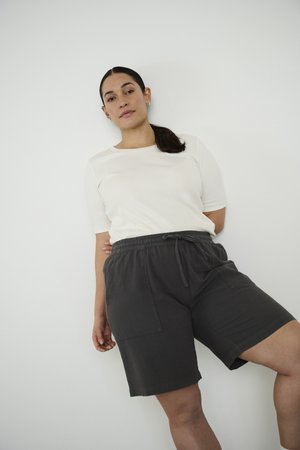 Kvinde med mørkt hår sat tilbage iført en hvid kortærmet skjorte og løse sorte shorts, der læner sig op ad en hvid væg.