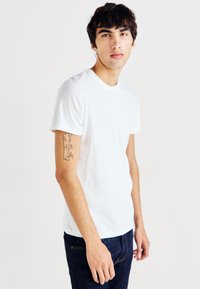 White cotton t-shirt with a crew neck and short sleeves, paired with dark blue jeans. Visible tattoo on the left forearm.