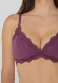 Femme portant un soutien-gorge en dentelle violet avec de fines bretelles réglables et des bords festonnés sur les bonnets et la bande sous la poitrine.