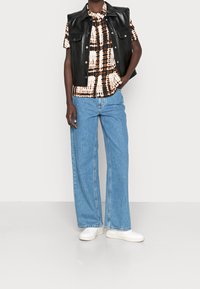 Un gilet en cuir noir sur une chemise tie-dye en crème et marron, associé à un jean large bleu clair et des baskets blanches.