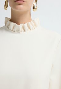 Top bianco con colletto arricciato, realizzato in tessuto morbido. Presenta grandi orecchini dorati, che sottolineano eleganza e design moderno.