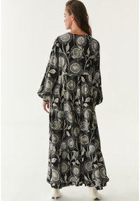 Langes schwarzes Kleid mit einem floralen Muster in Creme. Hat lange Ärmel und ein gestuftes Design, mit einem volantartigen Saum am unteren Ende.