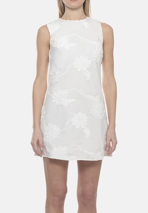 Donna che indossa un mini abito bianco senza maniche con ricamo floreale, in piedi davanti a uno sfondo chiaro e semplice.