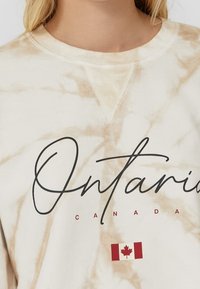 Crème-kleurig sweatshirt met een gemarmerd patroon. Bevat "Ontario" in zwarte schrift en een kleine Canadese vlag graphic. Getextureerde stof.