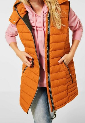 Kobieta ubrana w pomarańczową pikowaną kamizelkę bez rękawów na różowej bluzie z kapturem, ręce w kieszeniach kamizelki, w połączeniu z jasnoniebieskimi dżinsami.