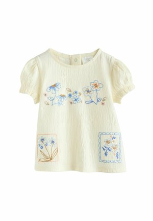 Ljusgul kortärmad topp med textilstruktur, prydd med blommig broderi i blått och orange på framsidan och två dekorativa lappar.