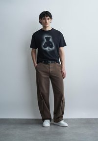 Jeune homme portant un t-shirt noir avec un motif ours, un pantalon cargo marron, des baskets blanches, debout contre un mur gris clair uni.