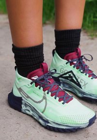 Chaussures de sport vertes et noires avec une tige en maille texturée, des lacets bordeaux et une semelle à motif. Présentent un logo 'GORE-TEX' sur le côté.