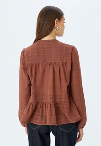 Blusa color ruggine in tessuto texturizzato, con orlo a peplo plissettato e maniche lunghe, dotata di un semplice scollo rotondo e motivo a quadri.