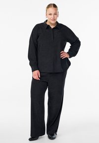 Schwarzes, abgestimmtes Outfit bestehend aus einem langärmligen Hemd mit Kragen und weiten Hosen. Beide haben einen glatten, strukturierten Stoff.