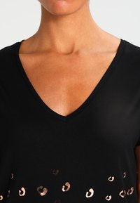 Černé tričko s výstřihem do V vyrobené z měkké látky, s metalickými leopardími skvrnami ve zlatých a měděných tónech blízko dolního lemu. Krátké rukávy.