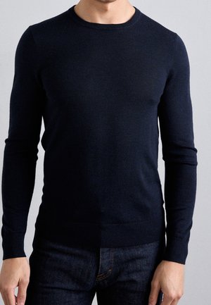 Homme portant un pull bleu marine ajusté à manches longues et un jean foncé devant un fond gris clair uni.