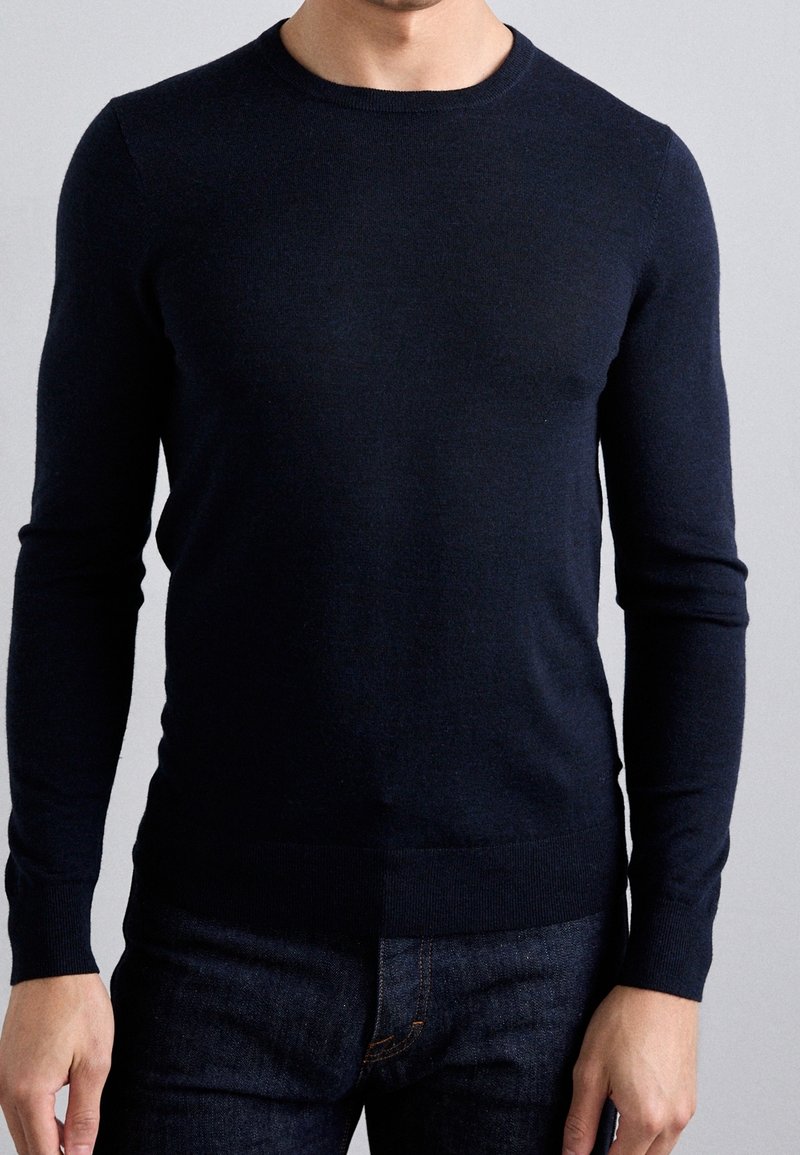 Homme portant un pull bleu marine ajusté à manches longues et un jean foncé devant un fond gris clair uni.