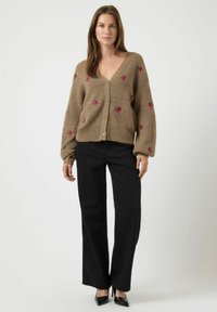 Cardigan marrone a maglia con scollatura a V, chiusura con bottoni e ricami floreali rosa. Abbinato a pantaloni neri a gamba larga e tacchi neri.