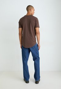 Tricou scurt din bumbac maro cu un tiv rotunjit, asortat cu blugi denim albaștri și pantofi negri, văzut din spate.