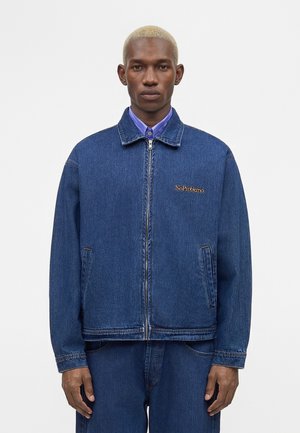 Mand med kort blondt hår iført en blå denimjakke og bukser, lilla skjorte synlig under jakkekraven.