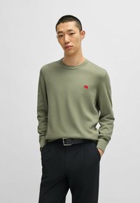 Langärmliger olivgrüner Pullover aus weichem Material mit Rundhalsausschnitt und rotem Markenpatch auf der Brust, kombiniert mit schwarzen Hosen.