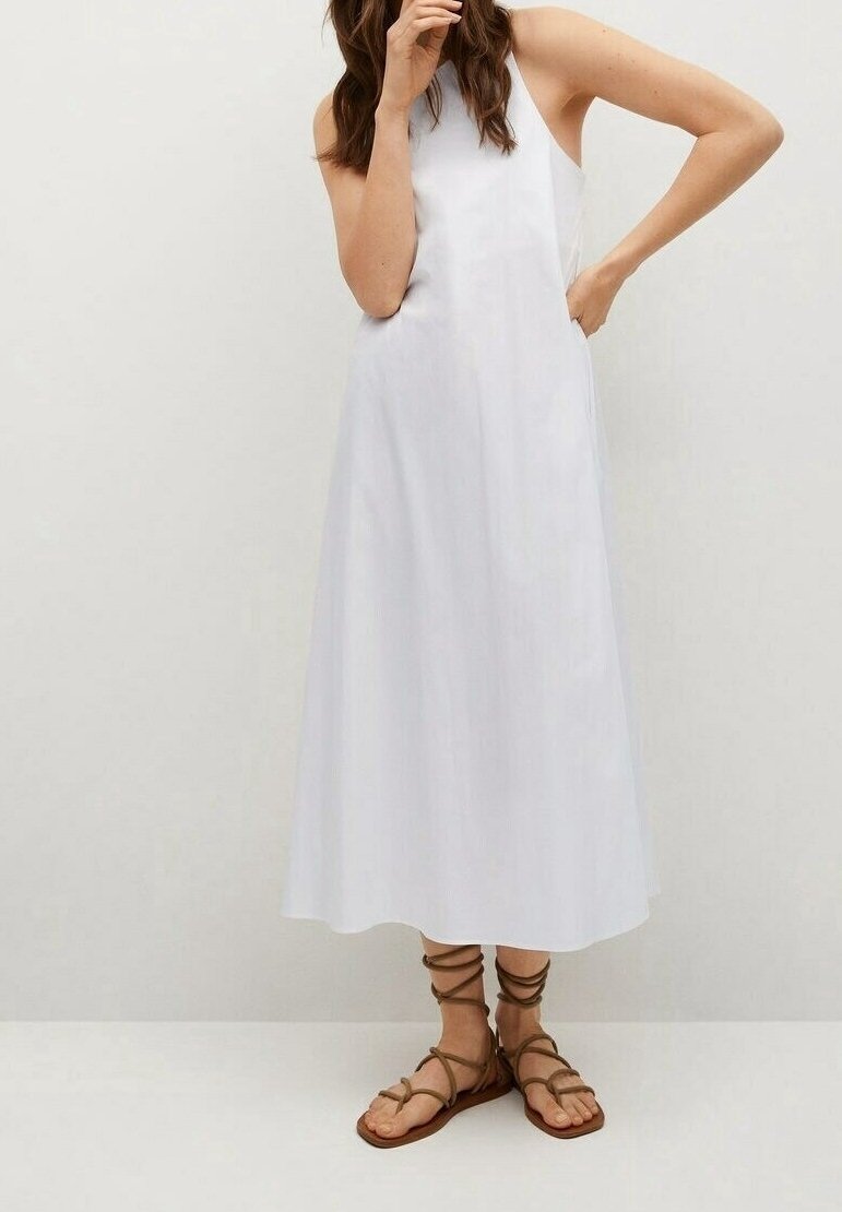 Femme en robe midi blanche sans manches et sandales plates marron à brides, debout devant un fond clair uni.