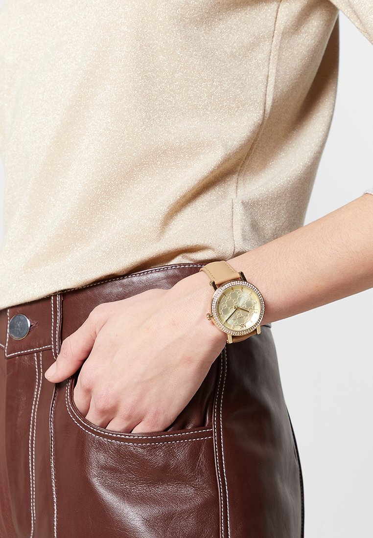 Reloj dorado con una cara redonda, pulsera de cuero ligera color beige y acentos de cristal alrededor del bisel. Lleva puesto en una mano contra un pantalón de cuero marrón.