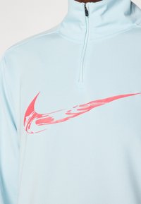 Jasnoniebieski polar z krótkim zamkiem wykonany z gładkiego materiału, z dużym różowym logo Nike na piersi. Posiada stojący kołnierz i detale szycia.