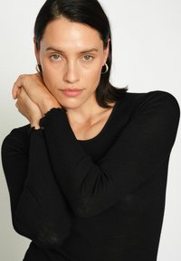 Fekete bordázott hosszúujjú felső, lekerekített nyakkivágással és fodros ujjvégekkel. A modellen semleges háttér és természetes bőrszín látható.