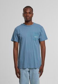 Helles blaues Baumwoll-T-Shirt mit kurzen Ärmeln, Rundhalsausschnitt und einer linken Brusttasche mit weißer Logo-Stickerei. Einfaches und lässiges Design.