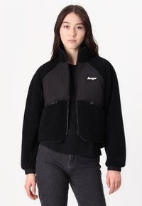Giacca cropped nera in morbido fleece con pannello nero a contrasto. Presenta tasche con zip e colletto alto. Accentuata con logo.