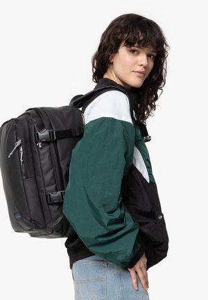 Schwarzer Rucksack mit mehreren Reißverschlussfächern, auf einer Schulter getragen. Der Träger trägt eine grün-schwarze Jacke mit glänzender Textur.