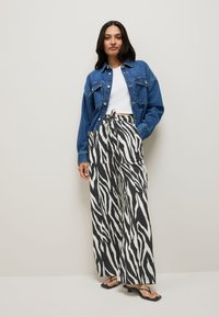 Woman wearing a blue denim jacket over a white crop top, black-and-white zebra print wide-leg pants, and black heeled sandals, standing against a plain wall.
