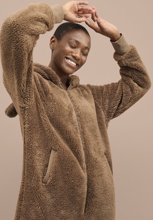 Lachend persoon met kort haar, gekleed in een bruine, harige jas met rits, die de armen boven het hoofd heft tegen een effen beige achtergrond.