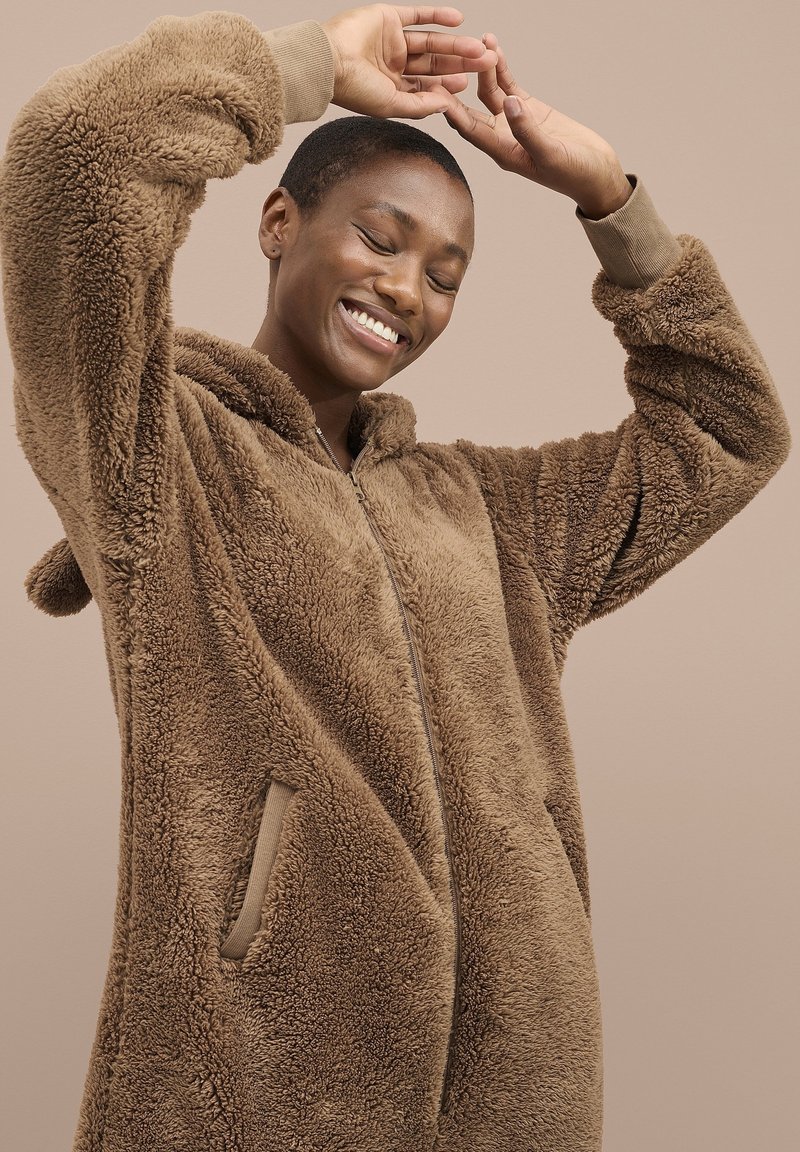 Uśmiechnięta osoba o krótkich włosach, ubrana w brązową, futrzaną kurtkę na zamek, unosząca ramiona nad głową na jednolitym beżowym tle.