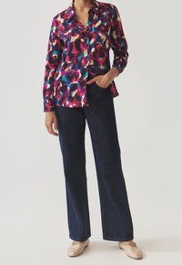 Blusa stampata colorata con motivi geometrici nelle tonalità di rosa, blu e beige. Abbinata a pantaloni ampi in denim scuro e scarpe basse beige.