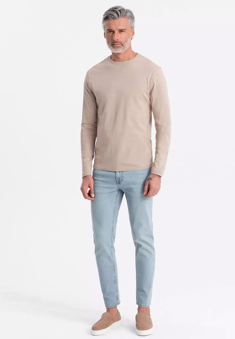 Vaaleanbeige pitkähihainen paita pyöreällä kaula-aukolla, yhdistettynä vaaleansinisiin slim fit -farkkuihin ja vaaleanbeigeihin liukujalkineisiin, neutraalia taustaa vasten.