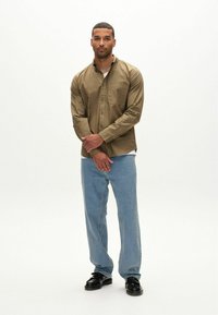 Olivgrünes Langarmhemd mit Knopfleiste, kombiniert mit hellblauen Jeans und schwarzen Schuhen. Einfache, strukturierte Passform und klare Linien.