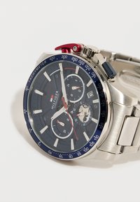 Ασημένιο μεταλλικό ρολόι χρονογράφου με μπλε στεφάνη ταχυμέτρου, μαύρο καντράν, τρεις υποκαντράν, κόκκινες λεπτομέρειες και ενωμένο μεταλλικό λουράκι σε λευκό φόντο.